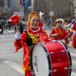 A woman in red and gold traditional attire carries and plays a large red bass drum during the Lunar New Year Parade in Chinatown, New York City.
