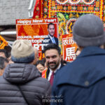 Mayor Zohran Mamdani smiles while speaking with attendees at the Lunar New Year Parade in Chinatown, with a red βHappy New Yearβ sign visible behind him.