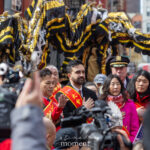 Mayor Zohran Mamdani stands with his hand over his heart during the Pledge of Allegiance at the Lunar New Year Parade in Chinatown, New York City, with a dragon costume behind him.