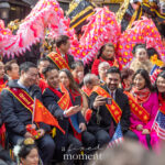 Mayor Zohran Mamdani holds a smartphone to take a group selfie with community leaders wearing red sashes during the Lunar New Year Parade in Chinatown, New York City, with pink dragon costumes in the background.