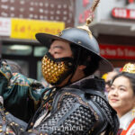 A parade participant wearing a wide-brimmed helmet, gold face covering, and black armor holds a flag during the Lunar New Year Parade in Chinatown, New York City.