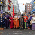 A group of parade participants wearing colorful traditional-style garments stand across a Chinatown street during the Lunar New Year Parade in New York City.