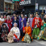 A large group of parade participants wearing colorful traditional Chinese-inspired garments pose together on a city street during the Lunar New Year Parade in Chinatown, New York City.