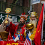 Two parade participants wearing ornate red and yellow traditional ceremonial costumes stand on a float during the Lunar New Year Parade in Chinatown, New York City.