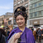 A participant wearing a purple traditional-style garment and ornate hair accessories stands in Chinatown during the Lunar New Year Parade in New York City.