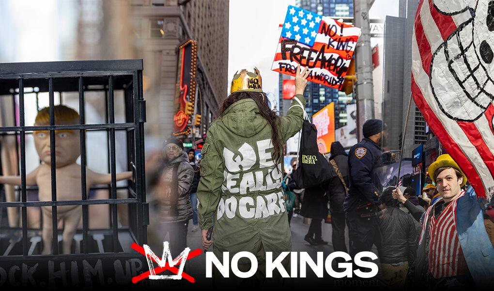 No Kings in New York: A Day of Protest, Presence, and a Measure of Hope A protester seen from behind wears a green jacket with the words “We Eat, Do Care” and holds a sign above their head in Times Square.