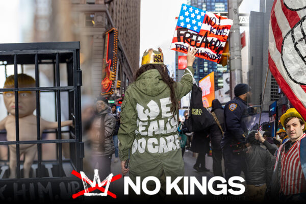 No Kings in New York: A Day of Protest, Presence, and a Measure of Hope A protester seen from behind wears a green jacket with the words “We Eat, Do Care” and holds a sign above their head in Times Square.