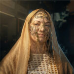 Cosplayer wearing a gold veil with chains and decorative face jewelry stands indoors during Awesome Con, with soft lighting highlighting the face and fabric details.