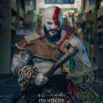 Cosplayer dressed as Kratos from God of War holds an axe while wearing body paint, leather armor, and chain props inside a convention center during Awesome Con.
