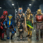 Group of cosplayers dressed as Mandalorian characters from Star Wars stand together on a convention floor wearing detailed armor in green, red, blue, gold, and silver during Awesome Con.