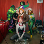 Group of cosplayers dressed as Marvel characters pose together at a convention booth, including She-Hulk, Rogue, and Spider-Gwen.
