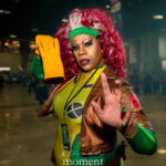 Cosplayer dressed as Rogue from X-Men poses with one hand extended toward the camera on the convention floor during Awesome Con, wearing a green and yellow bodysuit, brown jacket, and gloves.