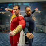 Two cosplayers dressed as Shazam and Superman stand back to back, extending their arms in a synchronized action pose in a convention hallway.