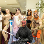 Group of cosplayers posing together, including a Darth Maul costume with a double-bladed lightsaber and several Jedi and fantasy-inspired outfits in a convention hallway.
