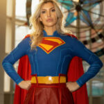 Cosplayer dressed as Supergirl from DC Comics stands with hands on hips inside a convention hall wearing a blue suit with a red cape and red skirt.