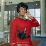 Cosplayer dressed as Uhura from Star Trek poses inside a convention center wearing a red Starfleet uniform with gold trim and holding a communicator, with a phaser in a waist holster.