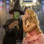 Cosplayer dressed as Glinda whispers to a cosplayer dressed as the Wicked Witch from The Wizard of Oz inside a convention center during Awesome Con, wearing a pink gown and black witch costume with green face makeup.