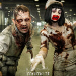 Two cosplayers in zombie-style makeup and blood-stained clothing reach toward the camera inside a convention hall.
