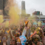 A large crowd raises their hands and throws colored powder into the air during the Holi Festival of Colors celebration at Pier 17 in New York City.