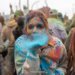 A participant covered in colorful powder blows blue powder from their hand while standing in a crowd during Holi at Pier 17 in New York City.