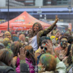 A participant covered in colorful powder raises a peace sign above a crowd celebrating the Holi Festival of Colors at Pier 17 in New York City.
