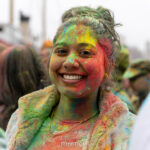 Dog wearing a harness with colorful powder on its face stands on the wooden deck during the Holi celebration at Pier 17 in New York City.