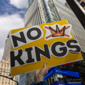 A hand holds a yellow sign reading “No Kings” with a crossed-out crown symbol against tall buildings in Manhattan.