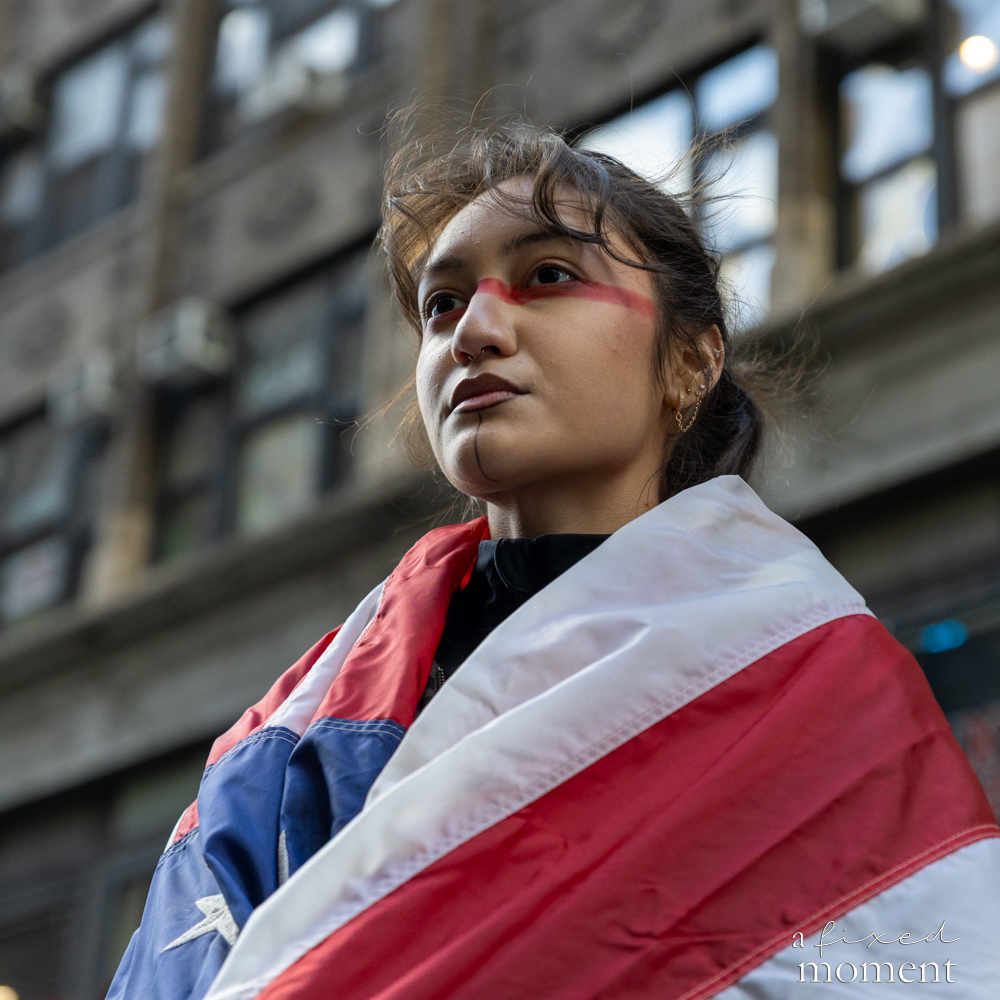 A protest participant with red face paint is wrapped in a flag during the No Kings protest in Manhattan.