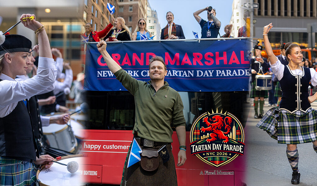 Sam Heughan stands in front of a Grand Marshal banner on a parade bus, raising his fist while holding a bottle of whisky as Highland dancers and drummers perform on Sixth Avenue in New York City.