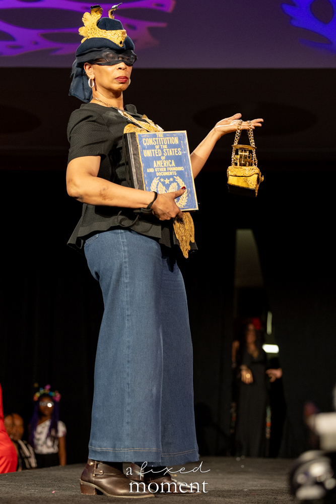 Participant carrying a Constitution book and gold handbag on the runway at Brooklyn's 2026 People's Ball
