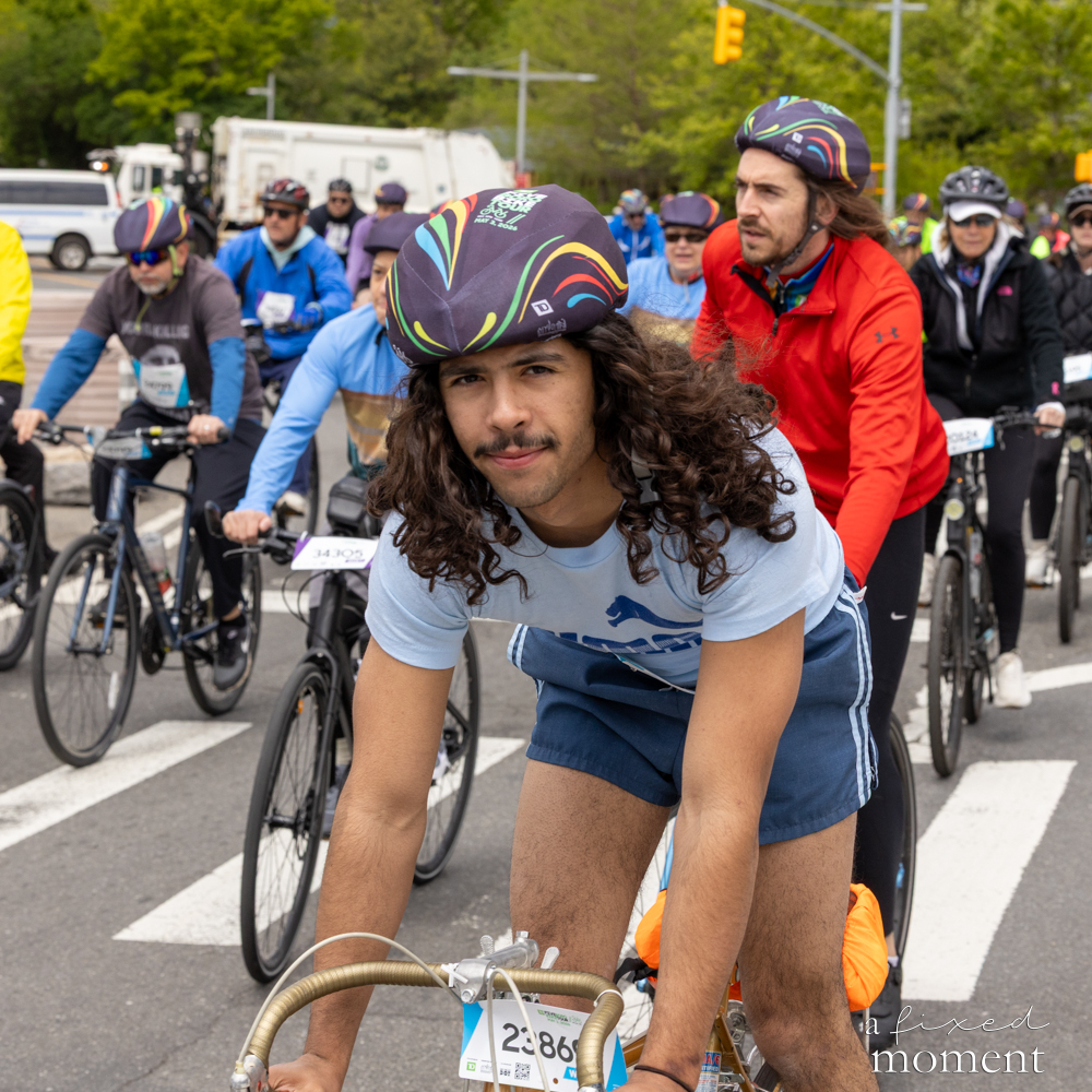 Long-haired cyclist rides through an intersection with a group at the Five Boro Bike Tour