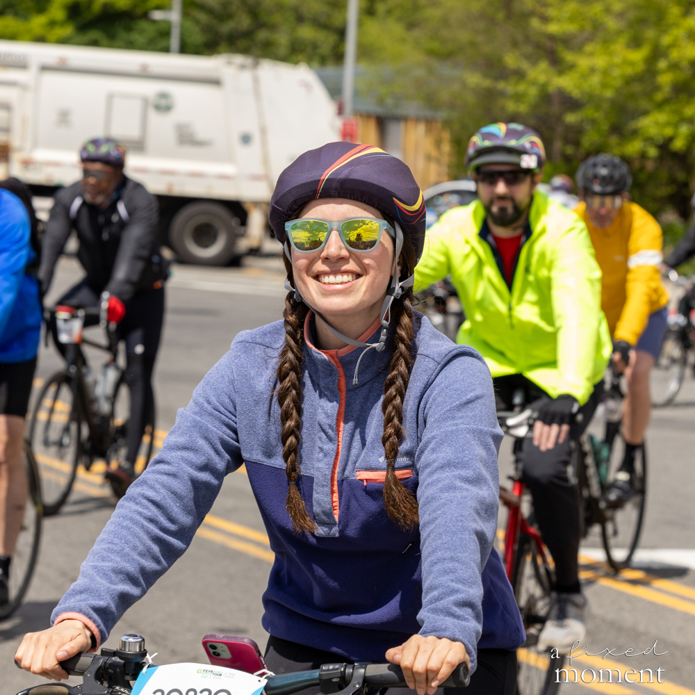 Smiling cyclist with braids and sunglasses rides with a pack at the Five Boro Bike Tour