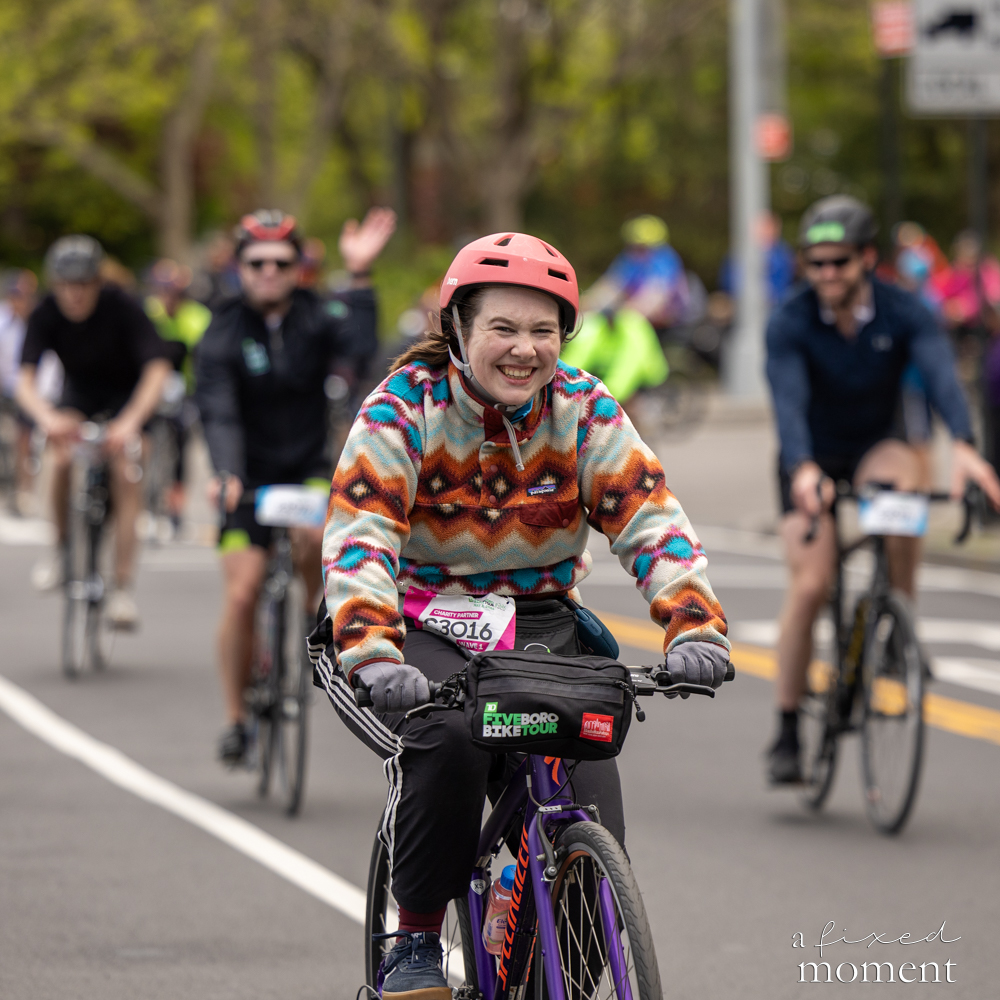 Smiling cyclist in a colorful sweater rides with a pack at the Five Boro Bike Tour