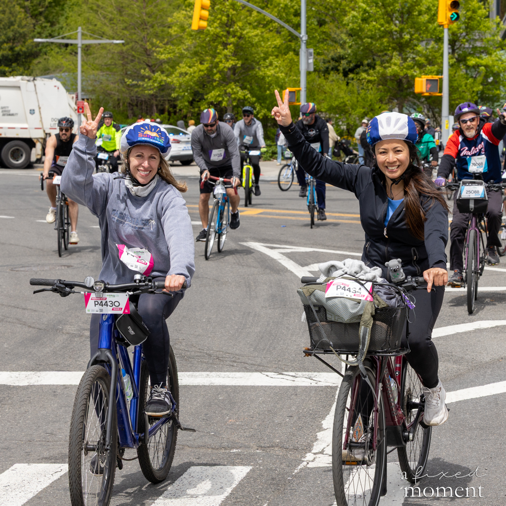 Two cyclists flash peace signs at an intersection during the 2026 Five Boro Bike Tour