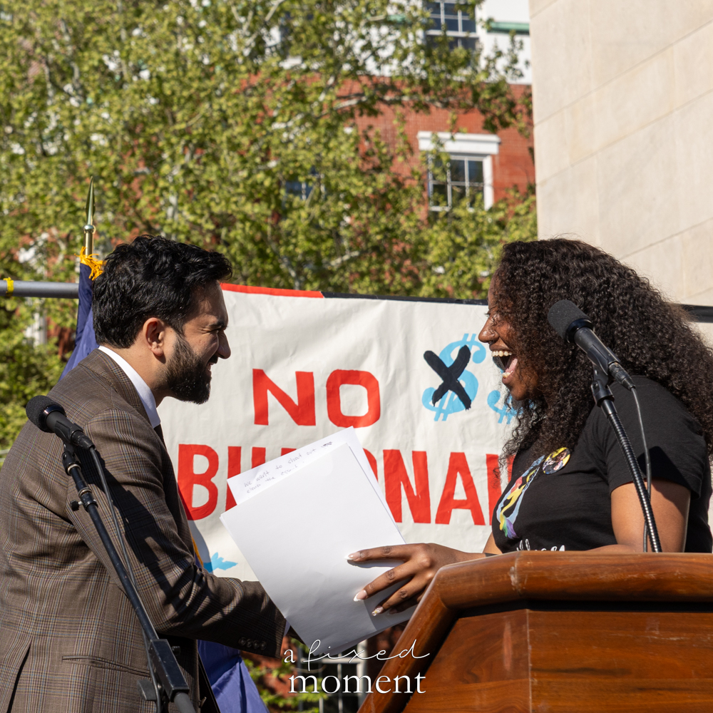 Mayor Zohran Mamdani speaks on stage near a No Billionaires banner