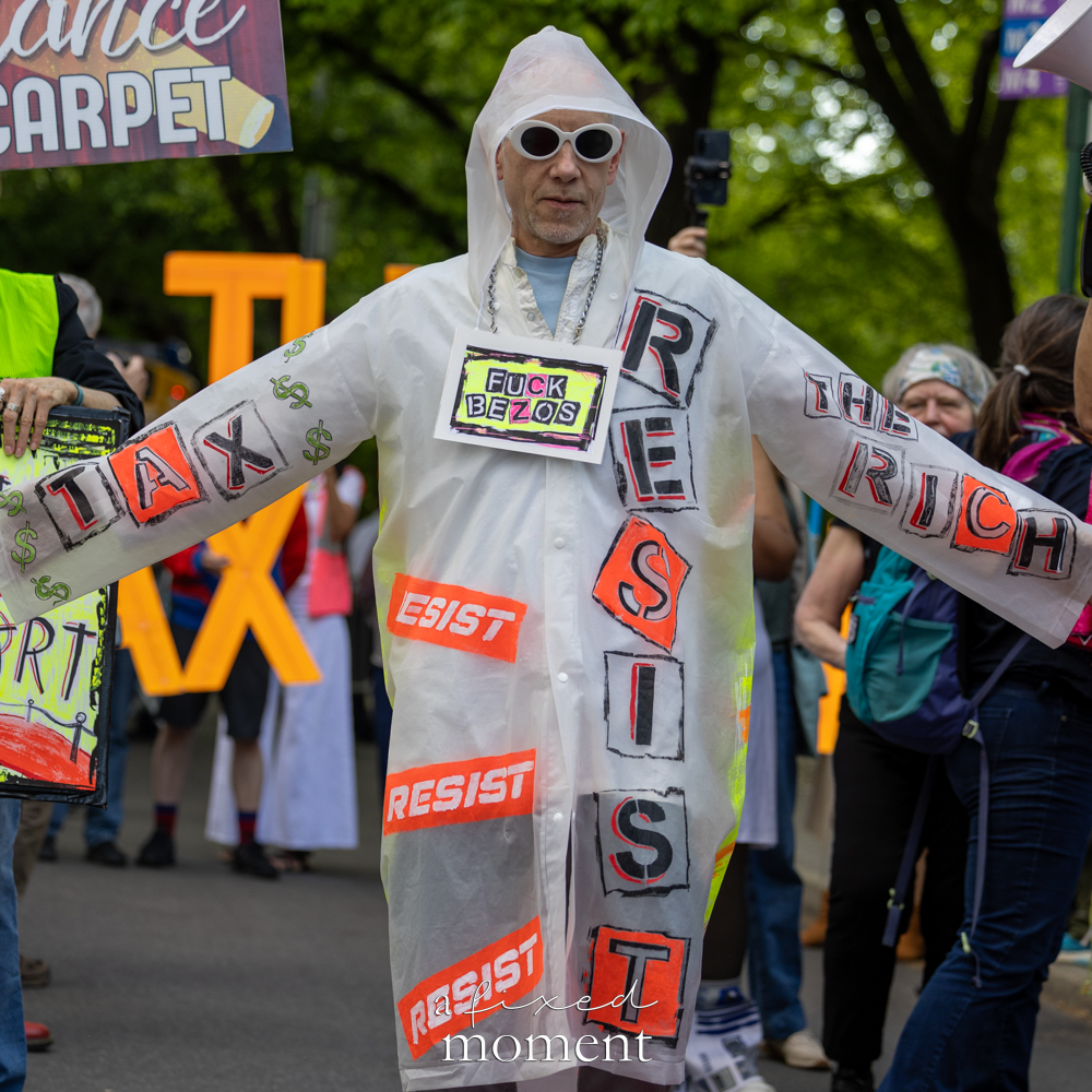 Costumed protester poses beneath signs reading Fuck Bezos, Tax, The Rich and Resist outside the Met Gala