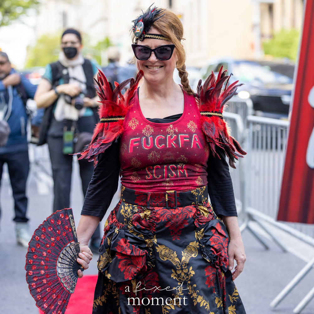 Participant in a red costume holds a fan with text reading Fuck Fascism outside the Met Gala
