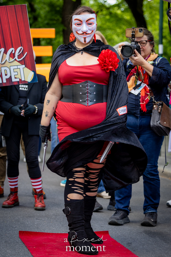 Masked participant in a red and black costume poses on the red carpet outside the Met Gala