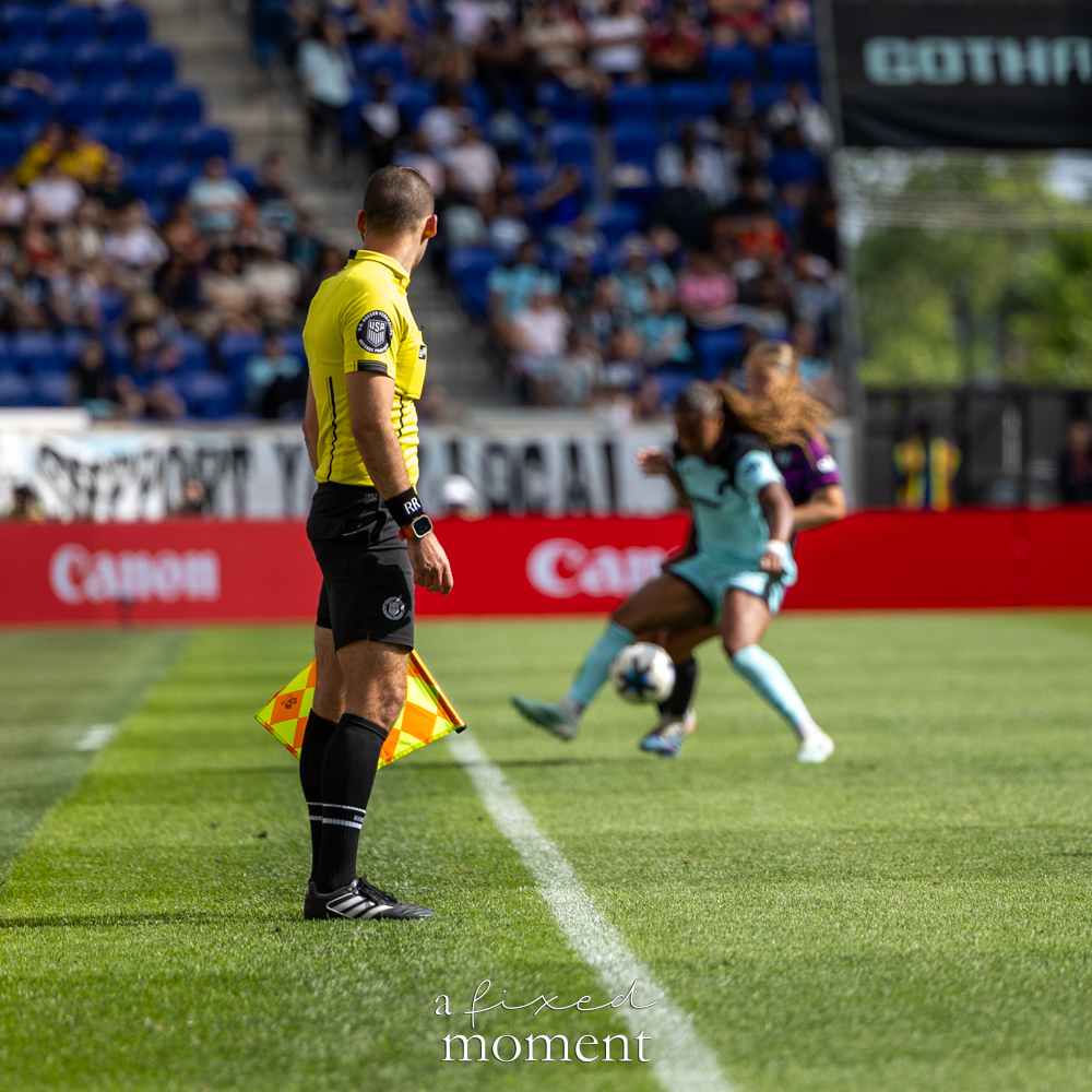 Assistant referee in a yellow shirt holding a checkered flag on the sideline while Gotham FC @gothamfc and Houston Dash @houstondash players compete in the blurred background.