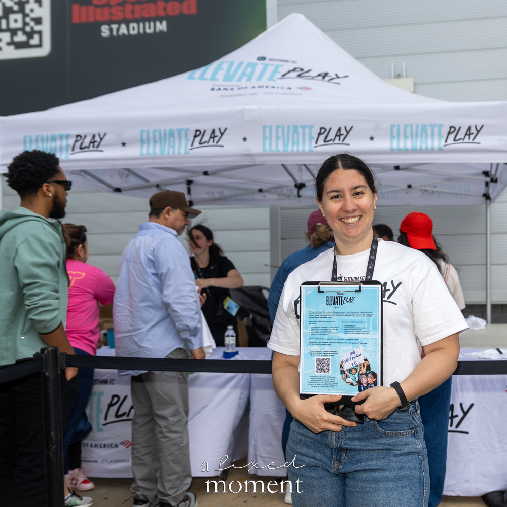 Smiling Gotham FC matchday representative holding an Elevate Play information sheet beneath a white branded tent outside Sports Illustrated Stadium.