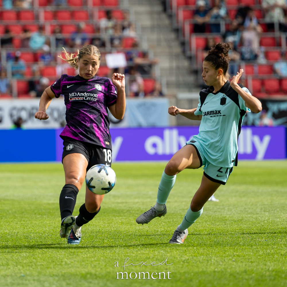 Gotham FC @gothamfc player in a light-blue kit challenging a Houston Dash @houstondash player in a black-and-purple kit as the ball moves between them.
