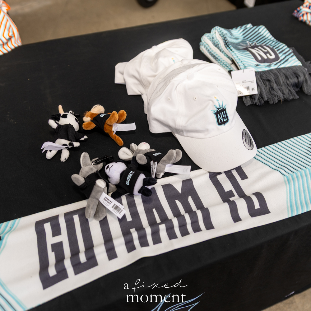 Gotham FC hats, plush toys and scarves arranged on a black merchandise table before a soccer match.