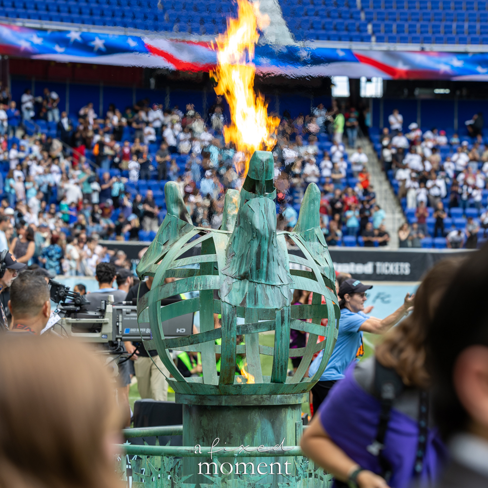 Gotham FC @gothamfc Statue of Liberty-style torch burning on the field with fans and blue stadium seats in the background.