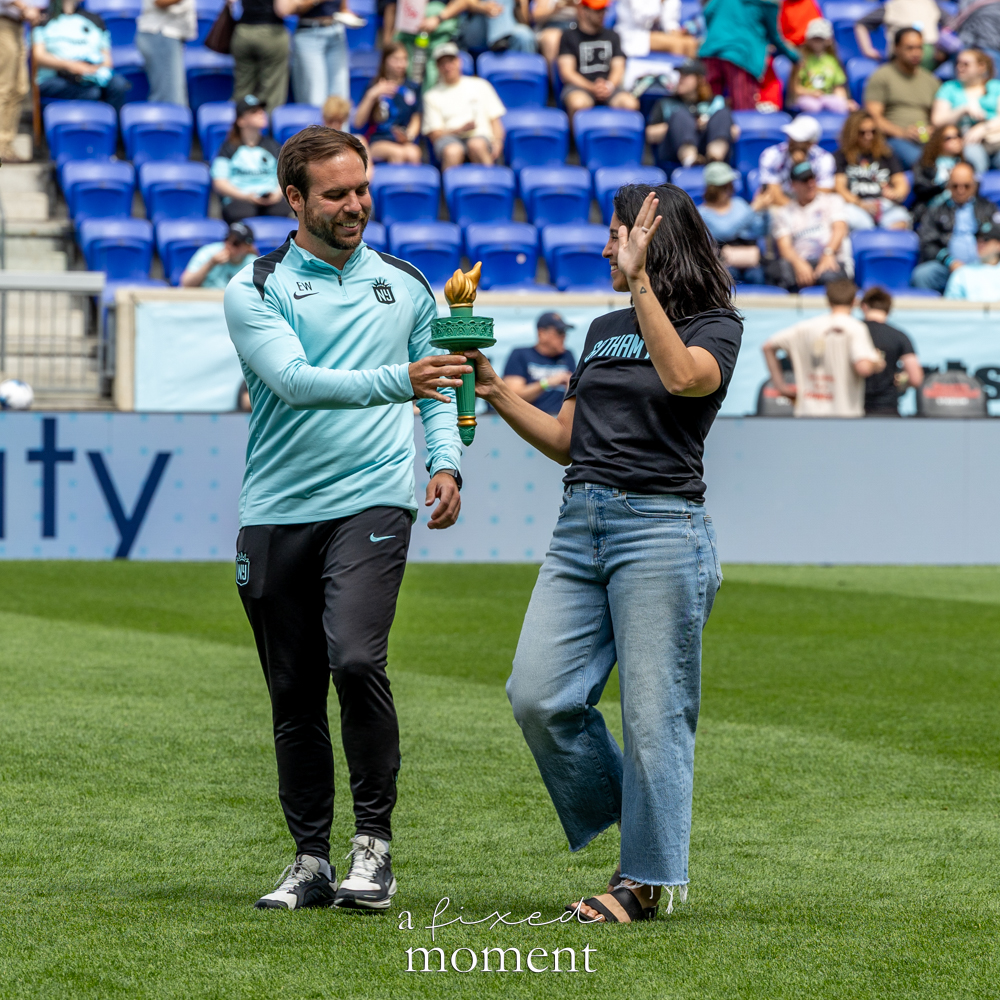 Gotham FC @gothamfc staff member handing a green torch prop to a supporter on the field with blue stadium seats behind them.
