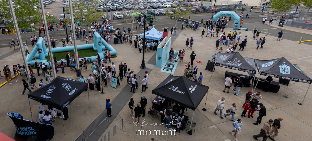 Wide overhead view of fans walking through the Gotham FC pregame plaza outside Sports Illustrated Stadium with tents, inflatable arches and team signage.