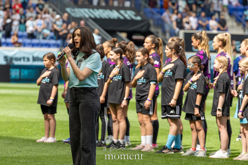 Singer holding a microphone on the soccer field while youth participants stand in a line with hands over their hearts behind her.
