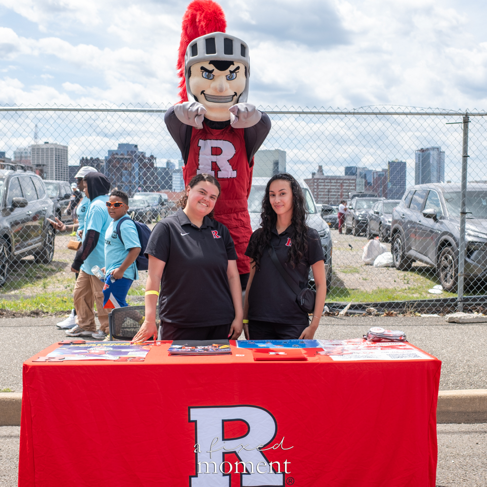 Two Rutgers representatives standing behind a red Rutgers table with the Scarlet Knight mascot behind them outside Sports Illustrated Stadium.