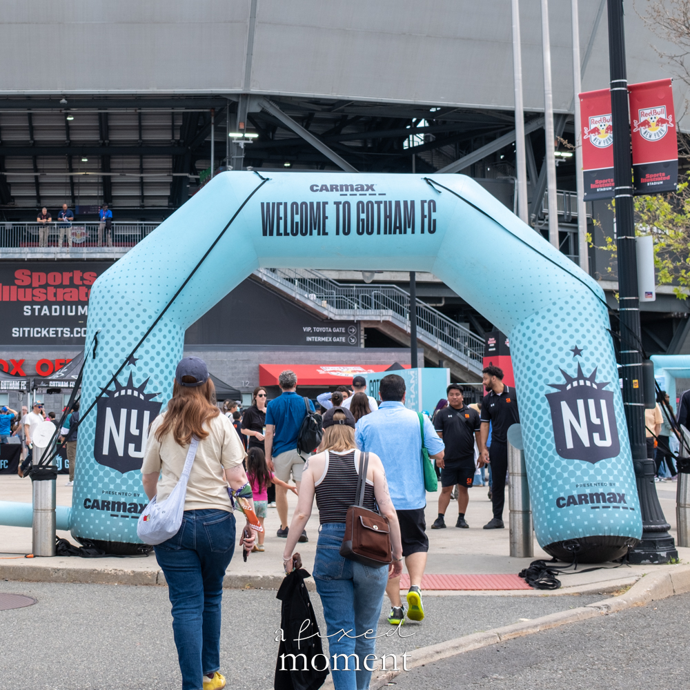 Fans walking toward Sports Illustrated Stadium beneath a light-blue Gotham FC welcome arch presented by CarMax.