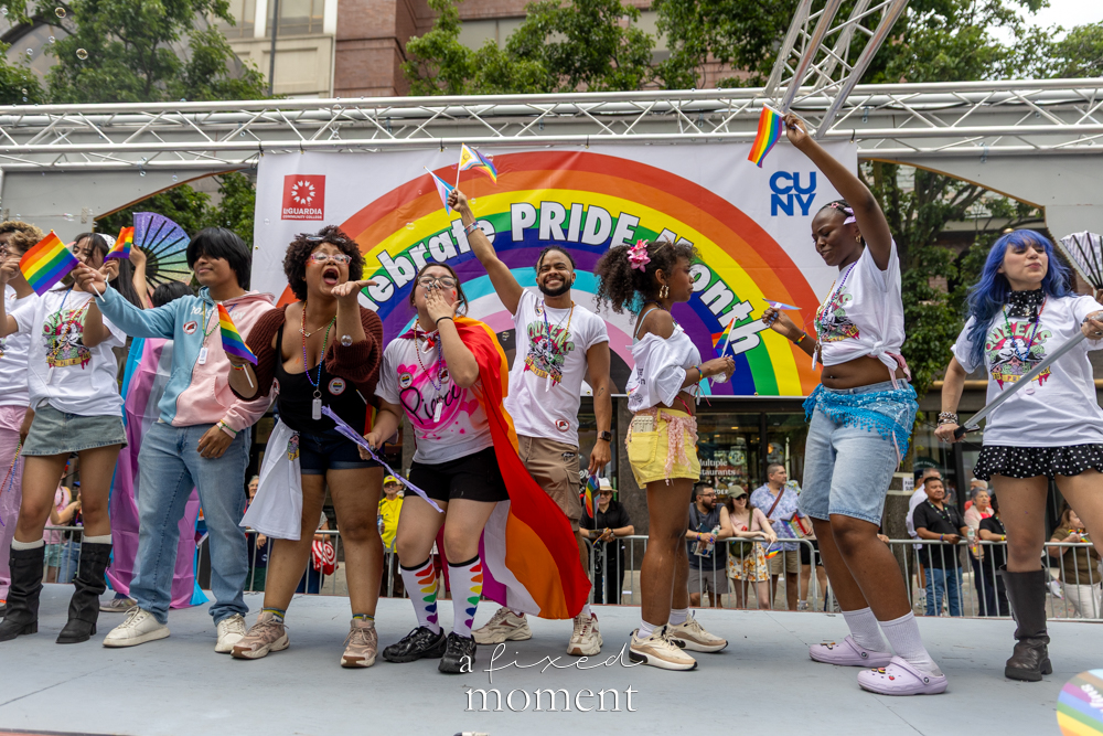 Performers appear onstage beneath an embrace Pride month backdrop.