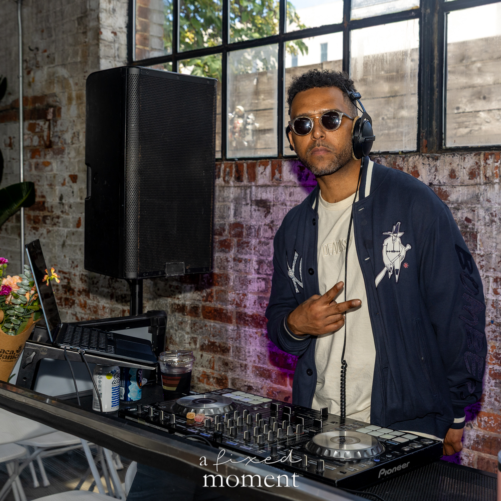 DJ wearing headphones and sunglasses stands behind a Pioneer DJ controller inside an industrial brick venue.